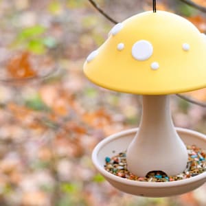 May include: A hanging bird feeder shaped like a mushroom with a bright yellow cap featuring white spots. The stem is a light beige color and connects to a circular tray base filled with a variety of small, colorful bird seeds and grains.