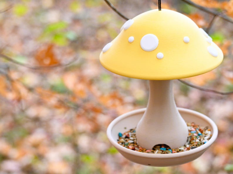 May include: A hanging bird feeder shaped like a mushroom with a bright yellow cap featuring white spots. The stem is a light beige color and connects to a circular tray base filled with a variety of small, colorful bird seeds and grains.