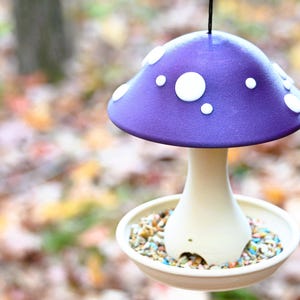 May include: A hanging bird feeder shaped like a mushroom with a purple cap featuring white circular spots. The stem is a light cream color and connects to a circular base tray filled with a variety of small, colorful bird seed pieces.