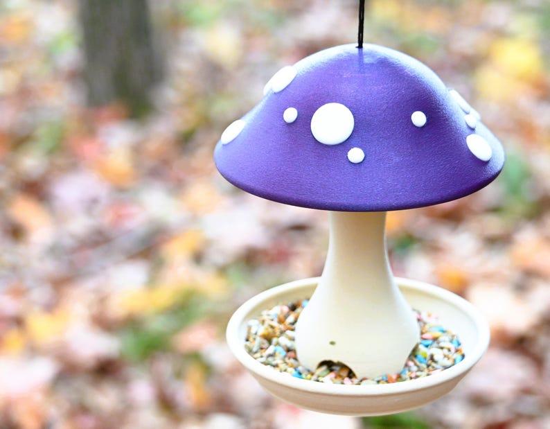 May include: A hanging bird feeder shaped like a mushroom with a purple cap featuring white circular spots. The stem is a light cream color and connects to a circular base tray filled with a variety of small, colorful bird seed pieces.