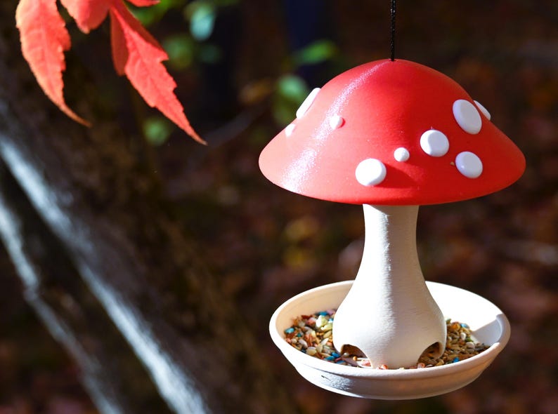 May include: A hanging bird feeder shaped like a mushroom with a bright red cap featuring white spots. The stem is off-white and supports a circular base filled with birdseed. The feeder is suspended by a thin black string from above.