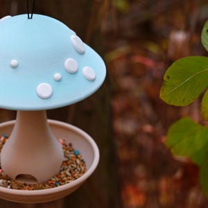 May include: A hanging bird feeder shaped like a mushroom with a light blue cap featuring white circular spots. The base is a tan, flared stem connected to a circular tray filled with a variety of bird seeds, including small colorful pieces.
