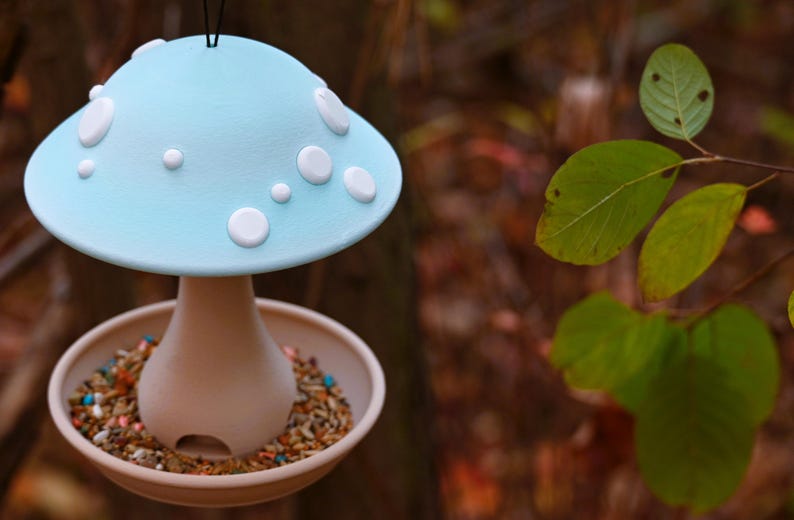 May include: A hanging bird feeder shaped like a mushroom with a light blue cap featuring white circular spots. The base is a tan, flared stem connected to a circular tray filled with a variety of bird seeds, including small colorful pieces.