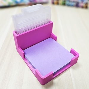 May include: A magenta desk organiser holding a stack of purple sticky notes. The organiser has a clear plastic top section and a rectangular base with raised edges. A practical and colourful office accessory for the desk.