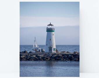 May include: A white lighthouse with a green trim stands on a rocky breakwater extending into the ocean. A sailboat with white sails is visible on the water to the left, and a person is riding a bicycle on the path near the lighthouse base.