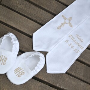 Baptism crib shoes and stole on a wood background.  White crib shoes are monogramed in champagne gold, rounded font - NKE.  White stole is embroidered in champagne gold with a scroll cross on one side and Nelle Elizabeth, 6-14-15 on the other side.