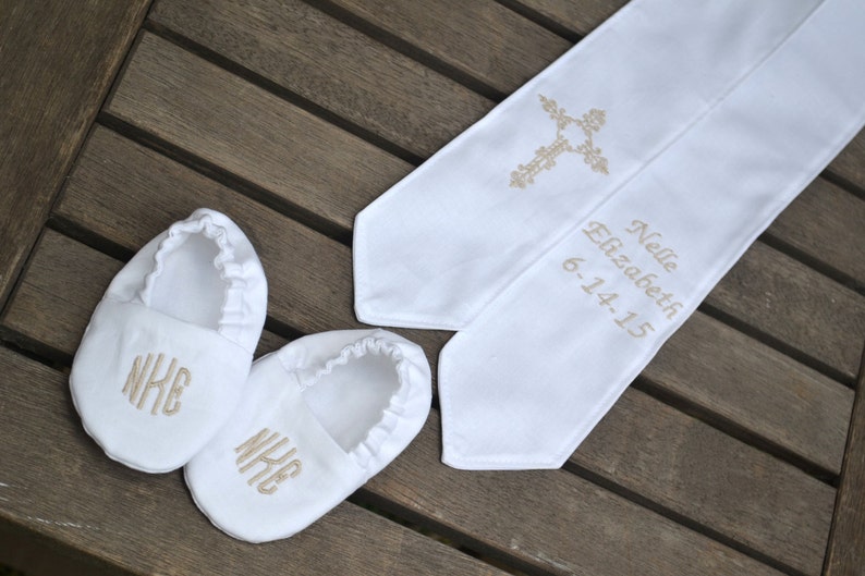 Baptism crib shoes and stole on a wood background.  White crib shoes are monogramed in champagne gold, rounded font - NKE.  White stole is embroidered in champagne gold with a scroll cross on one side and Nelle Elizabeth, 6-14-15 on the other side.