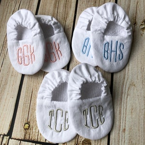 Wood background with 3 pairs of white crib shoes.  Top left with pink embroidery GGK in rounded font, top right with light blue embroidery BHS, bottom with silver grey embroidery TCE.