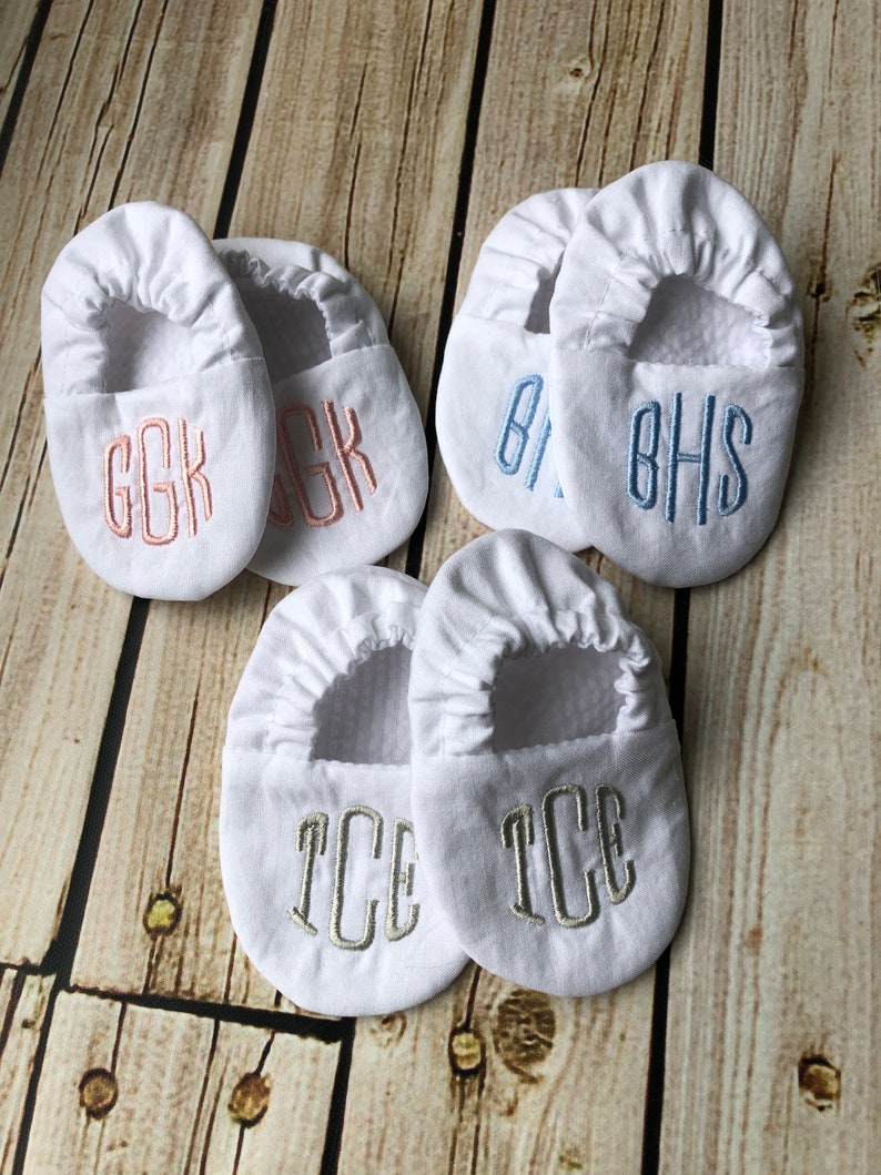 Wood background with 3 pairs of white crib shoes.  Top left with pink embroidery GGK in rounded font, top right with light blue embroidery BHS, bottom with silver grey embroidery TCE.