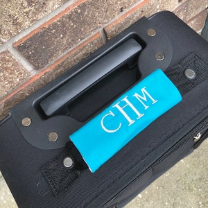 May include: A black suitcase with a turquoise luggage handle strap with the monogram "CHM" embroidered in white.