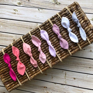 May include: A collection of seven fabric headbands in various colors and patterns, including solid pinks, a polka dot, and striped designs. The headbands are arranged on a woven basket with a light wood grain background.