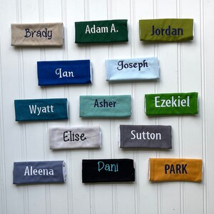 May include: A collection of thirteen rectangular fabric bands in various colors including beige, dark green, olive, blue, light blue, teal, sage, lime green, gray, charcoal, black, and mustard, each embroidered with a unique name in contrasting thread.