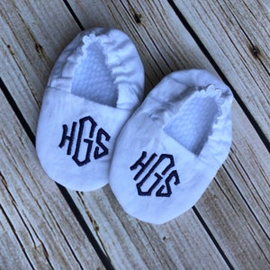 White crib shoes on a wood background.  Crib shoes are monogramed in navy blue, diamond font - HGS