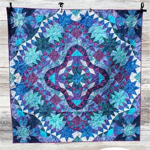 May include: A quilted wall hanging with a geometric starburst pattern in shades of blue, purple, and teal. The quilt features the text "Falling from the Stars" and is designed by Amanda Bauer. The quilt is displayed on a wooden background.