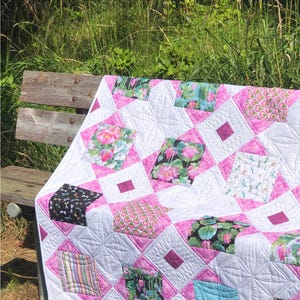 May include: A handmade quilt with a white background and pink diamond-shaped accents. The quilt features various patterned squares with floral and geometric designs. The quilt is draped over a wooden bench outdoors.