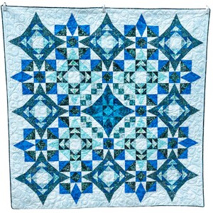May include: A large, square quilt with a geometric pattern in shades of blue and teal. The design features diamond and square shapes, creating a symmetrical and visually appealing textile. The quilt is hanging against a white background.