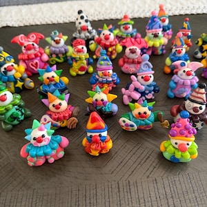 Puede incluir: Una colección de pequeñas figuras de payasos coloridas hechas de arcilla. Cada figura presenta un atuendo, sombrero y detalles faciales pintados únicos, incluyendo narices rojas y expresiones variadas en una amplia gama de colores brillantes.
