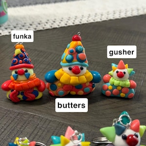 May include: A collection of colourful clown figurines made of clay, featuring various patterns and designs. The figurines are labelled with the names funka, butters, and gusher. Each clown has a red nose, a pointed hat, and a ruffled collar.