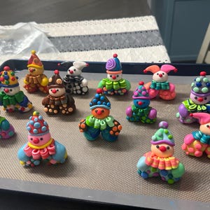May include: A collection of thirteen small, colourful clown figurines crafted from polymer clay. Each figure features a unique combination of bright colours, including polka-dot hats, ruffled collars, and rounded bodies, arranged on a baking sheet.