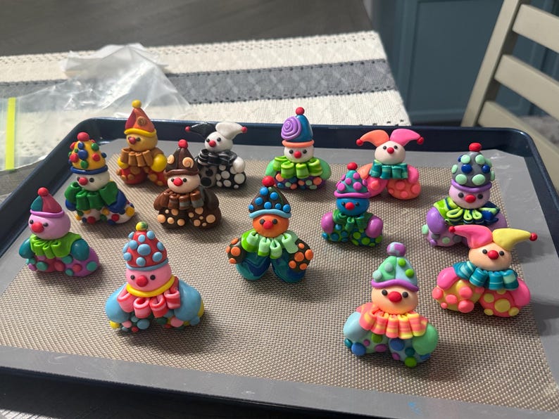 May include: A collection of thirteen small, colourful clown figurines crafted from polymer clay. Each figure features a unique combination of bright colours, including polka-dot hats, ruffled collars, and rounded bodies, arranged on a baking sheet.