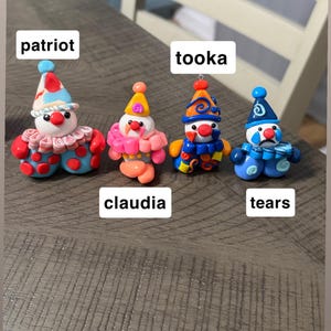 May include: Four small clown figurines made of clay. The first, labelled patriot, has a red, white, and blue hat and outfit. The second, claudia, is pink and orange. The third, tooka, is blue and orange. The fourth, tears, is blue with a sad face.