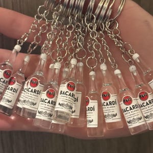 Puede incluir: Un conjunto de llaveros en miniatura con botellas de Bacardi Carta Blanca. Cada botella transparente tiene una etiqueta blanca con texto rojo y negro y un llavero plateado.