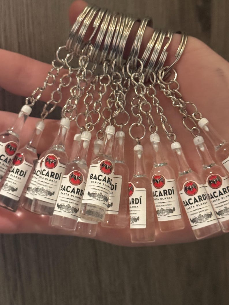 Puede incluir: Un conjunto de llaveros en miniatura con botellas de Bacardi Carta Blanca. Cada botella transparente tiene una etiqueta blanca con texto rojo y negro y un llavero plateado.