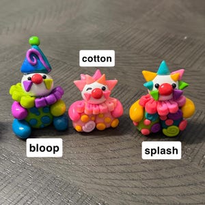 May include: Three small clown figurines made of colourful clay. The left figure is labelled bloop, wearing a blue hat and green and purple ruff. The centre figure is labelled cotton, in shades of pink and orange. The right figure is labelled splash.