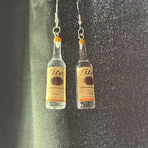 Puede incluir: Un par de pendientes artesanales con forma de botellas de vodka Tito's. Las botellas en miniatura tienen una etiqueta amarilla con el nombre de la marca y texto. Los pendientes tienen ganchos plateados.