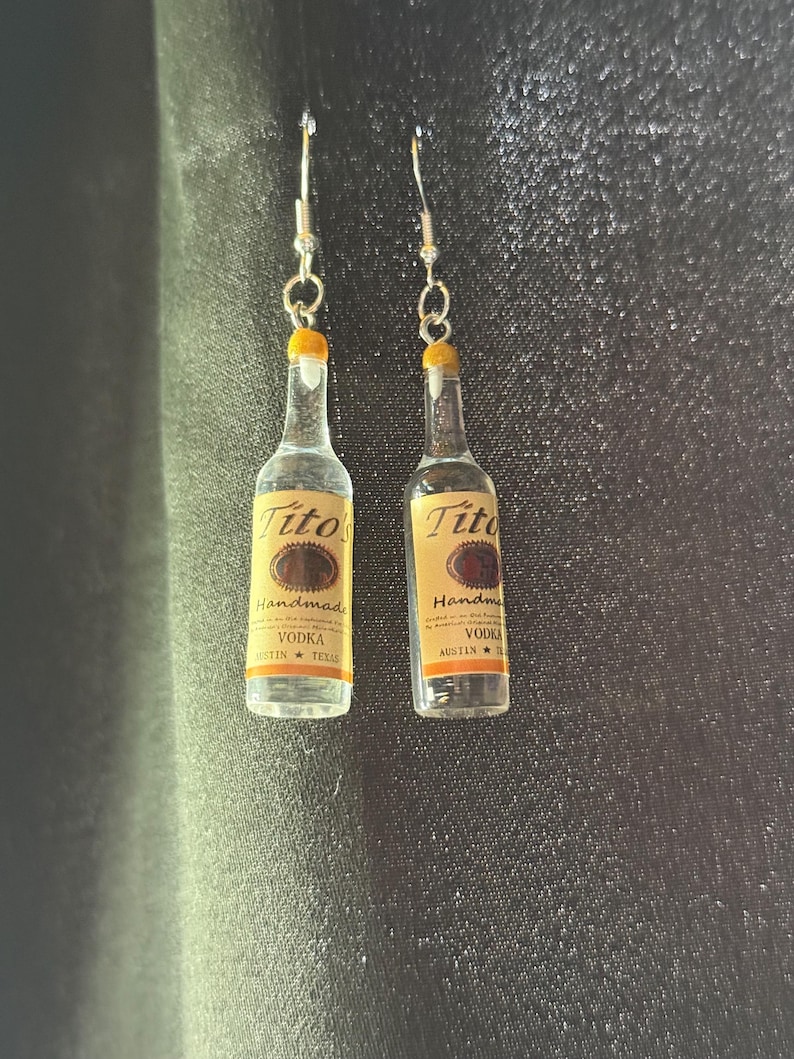 Puede incluir: Un par de pendientes artesanales con forma de botellas de vodka Tito's. Las botellas en miniatura tienen una etiqueta amarilla con el nombre de la marca y texto. Los pendientes tienen ganchos plateados.