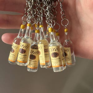 Puede incluir: Un conjunto de llaveros en miniatura con forma de botellas de vodka Tito's. Cada botella transparente tiene una tapa amarilla y una etiqueta con el nombre de la marca. Los llaveros est&aacute;n unidos a anillos de metal plateado.