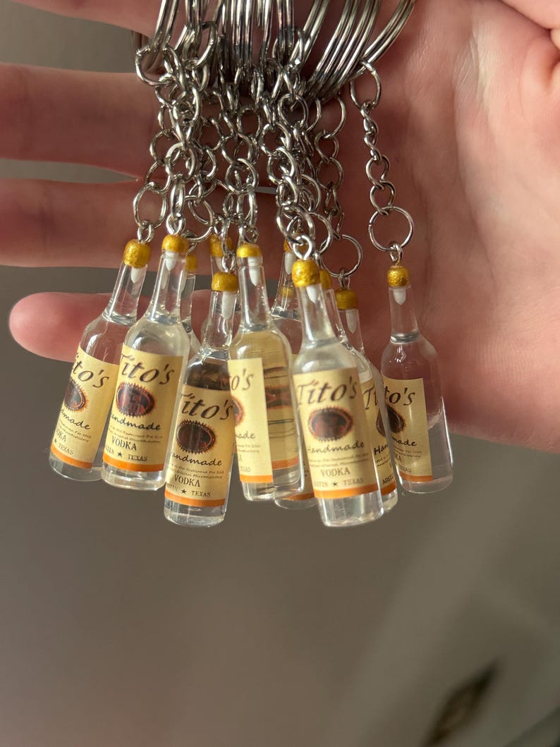 Puede incluir: Un conjunto de llaveros en miniatura con forma de botellas de vodka Tito's. Cada botella transparente tiene una tapa amarilla y una etiqueta con el nombre de la marca. Los llaveros est&aacute;n unidos a anillos de metal plateado.