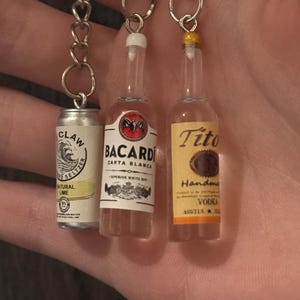 Puede incluir: Llaveros en miniatura con una lata de seltzer White Claw, una botella de Bacardi Carta Blanca y una botella de Tito's Handmade Vodka. Cada llavero est&aacute; unido a una cadena plateada. La lata de seltzer es plateada y blanca, mientras que las botellas son transparentes con etiquetas coloridas.