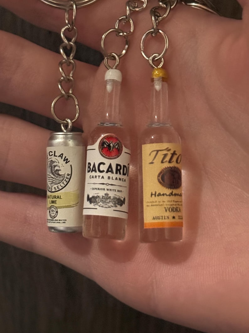 Puede incluir: Llaveros en miniatura con una lata de seltzer White Claw, una botella de Bacardi Carta Blanca y una botella de Tito's Handmade Vodka. Cada llavero est&aacute; unido a una cadena plateada. La lata de seltzer es plateada y blanca, mientras que las botellas son transparentes con etiquetas coloridas.
