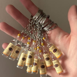 Puede incluir: Llaveros en miniatura de botellas de vodka Tito's Handmade. Cada botella transparente tiene una etiqueta amarilla y marr&oacute;n con el nombre de la marca. Los llaveros tienen anillos y cadenas de metal plateado.