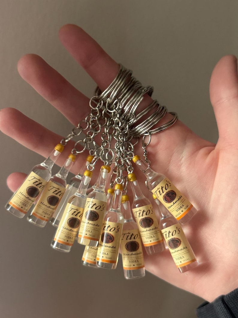 Puede incluir: Llaveros en miniatura de botellas de vodka Tito's Handmade. Cada botella transparente tiene una etiqueta amarilla y marr&oacute;n con el nombre de la marca. Los llaveros tienen anillos y cadenas de metal plateado.