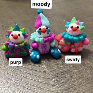 May include: Three small clown figurines made of colourful clay. The left one is labelled purp and has purple, green, and blue patterns. The centre one is labelled moody and features blue, purple, and pink colours. The right one is labelled swirly.
