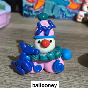 May include: A small clay figurine of a clown with a white face, red nose, and pink hat adorned with a blue balloon animal. The clown wears a teal and white striped ruffled collar and holds a blue balloon animal. The text ballooney is visible.