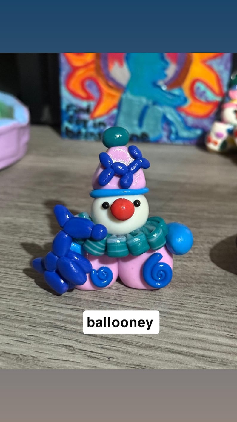 May include: A small clay figurine of a clown with a white face, red nose, and pink hat adorned with a blue balloon animal. The clown wears a teal and white striped ruffled collar and holds a blue balloon animal. The text ballooney is visible.