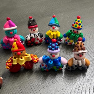 May include: A collection of seven small, handcrafted clay clown figurines. Each figure features a unique combination of vibrant colours, including polka-dot patterns, ruffled collars, and pointed hats. The clowns have round noses and expressive faces.