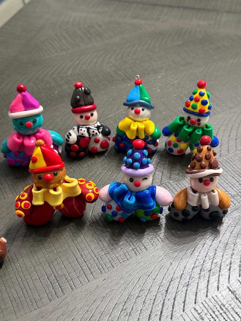 May include: A collection of seven small, handcrafted clay clown figurines. Each figure features a unique combination of vibrant colours, including polka-dot patterns, ruffled collars, and pointed hats. The clowns have round noses and expressive faces.