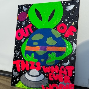 May include: A colorful painting depicts a green alien figure holding a blue and green planet. The artwork features the words "OUT OF THIS WORLD" in bright pink, set against a black starry background. A UFO is also present.