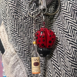 Puede incluir: Un llavero con un dije de mariquita roja con lunares negros y una botella de Vodka Tito's en miniatura. El dije de mariquita est&aacute; unido a una cadena plateada y un llavero. La botella tiene una tapa dorada.