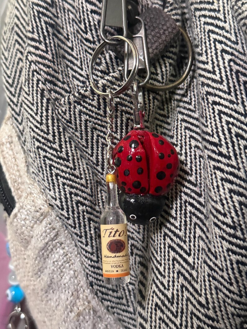 Puede incluir: Un llavero con un dije de mariquita roja con lunares negros y una botella de Vodka Tito's en miniatura. El dije de mariquita est&aacute; unido a una cadena plateada y un llavero. La botella tiene una tapa dorada.
