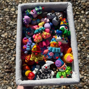 May include: A rectangular fabric basket filled with a collection of small, colourful clown figurines made of polymer clay. The clowns feature various bright colours, including red, blue, yellow, green, and purple, with distinct hats, ruffles, and polka-dot patterns.
