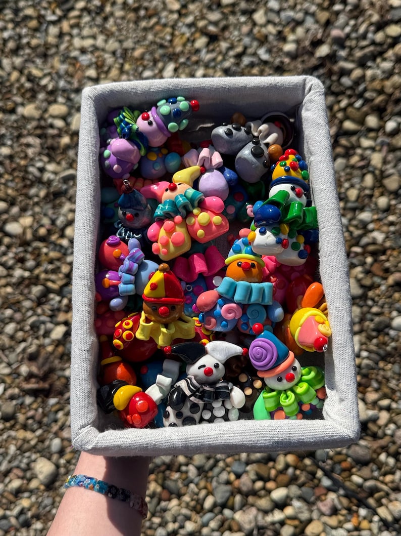May include: A rectangular fabric basket filled with a collection of small, colourful clown figurines made of polymer clay. The clowns feature various bright colours, including red, blue, yellow, green, and purple, with distinct hats, ruffles, and polka-dot patterns.