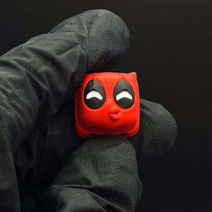Puede incluir: Una tecla Deadpool roja y cuadrada con ojos blancos y una boca pequeña y fruncida. La tecla se sostiene en una mano enguantada de negro sobre un fondo oscuro. Un artículo de colección para entusiastas de teclados mecánicos.