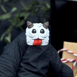 May include: A white and grey figurine with a red tongue and brown horns. The figurine is held by a gloved hand. A small gift box with a red and white string and a gold bell is in the background. A black candle is also in the background.