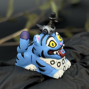 May include: A blue and black figurine of a cartoon tiger with yellow eyes, a red tongue, and a small hat. The tiger is lying down with its tail raised. The figurine is on a black surface.