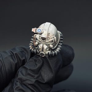Op de afbeelding: Een zilverkleurige ring in de vorm van een futuristische helm, met ingewikkelde details zoals een vizier, ademhalingsapparaat en een cirkelvormig blauw accent. De ring wordt vastgehouden door een gehandschoende hand tegen een donkere achtergrond.